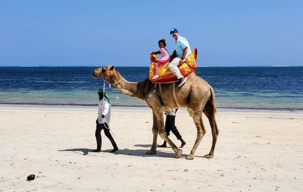 Jason Sucher and Bella Van Leeuwen riding a camel on the beach.